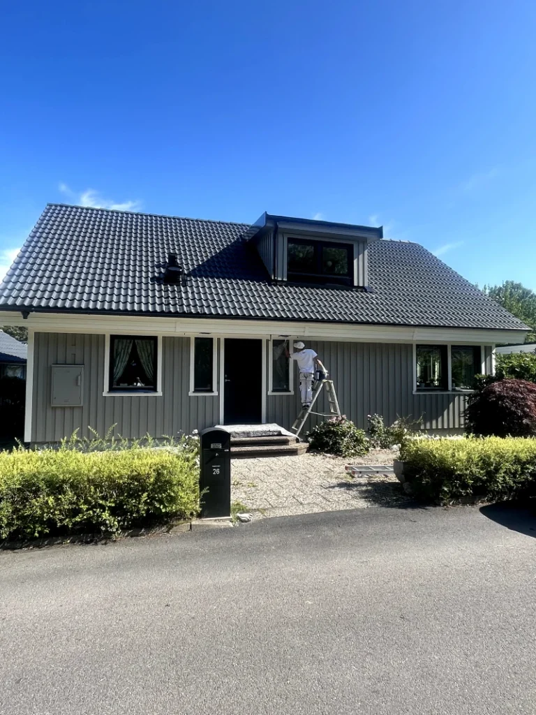 Fasadmålare applicerar ljusgrå färg på villa i solsken, förbättrar estetik och skydd mot väder