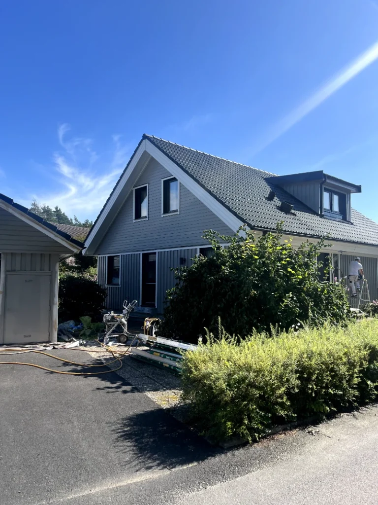 Målare i arbete med att skrapa och förbereda en villa för fasadmålning i dagsljus, omgiven av byggställningar.